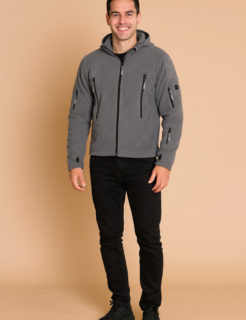 Grey softshell jacket with hood, multiple zippered pockets, and long sleeves with thumbholes, styled with black jeans and sneakers.