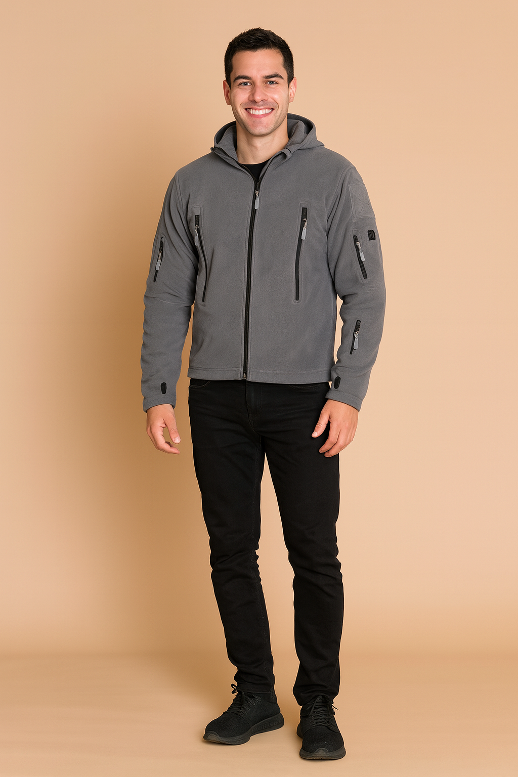 Grey softshell jacket with hood, multiple zippered pockets, and long sleeves with thumbholes, styled with black jeans and sneakers.