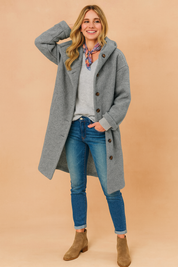 Long grey wool coat with button closure, paired with a light grey V-neck sweater, slim blue jeans, and taupe suede ankle boots.