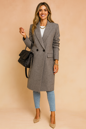 Elegant long grey wool double-breasted coat with two black buttons, structured lapels, tailored cut, and classic mid-length design, styled with light blue skinny jeans, beige suede pumps, a black quilted handbag with chain strap, and gold accessories.