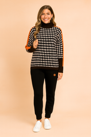 Oversized houndstooth turtleneck sweater with bold orange stripe detailing on sleeves, paired with black jogger pants and white sneakers.