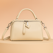 Ivory leather handbag with top handle, silver zipper, leather tag detail, and detachable shoulder strap on a warm gradient platform.