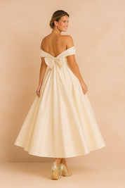 Elegant ivory off-the-shoulder evening gown with oversized back bow detail, voluminous A-line skirt, and paired with shimmering gold high heels.