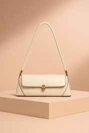 Ivory leather shoulder bag with gold clasp and long strap, displayed on a beige pedestal against a warm sand-to-blush gradient background.