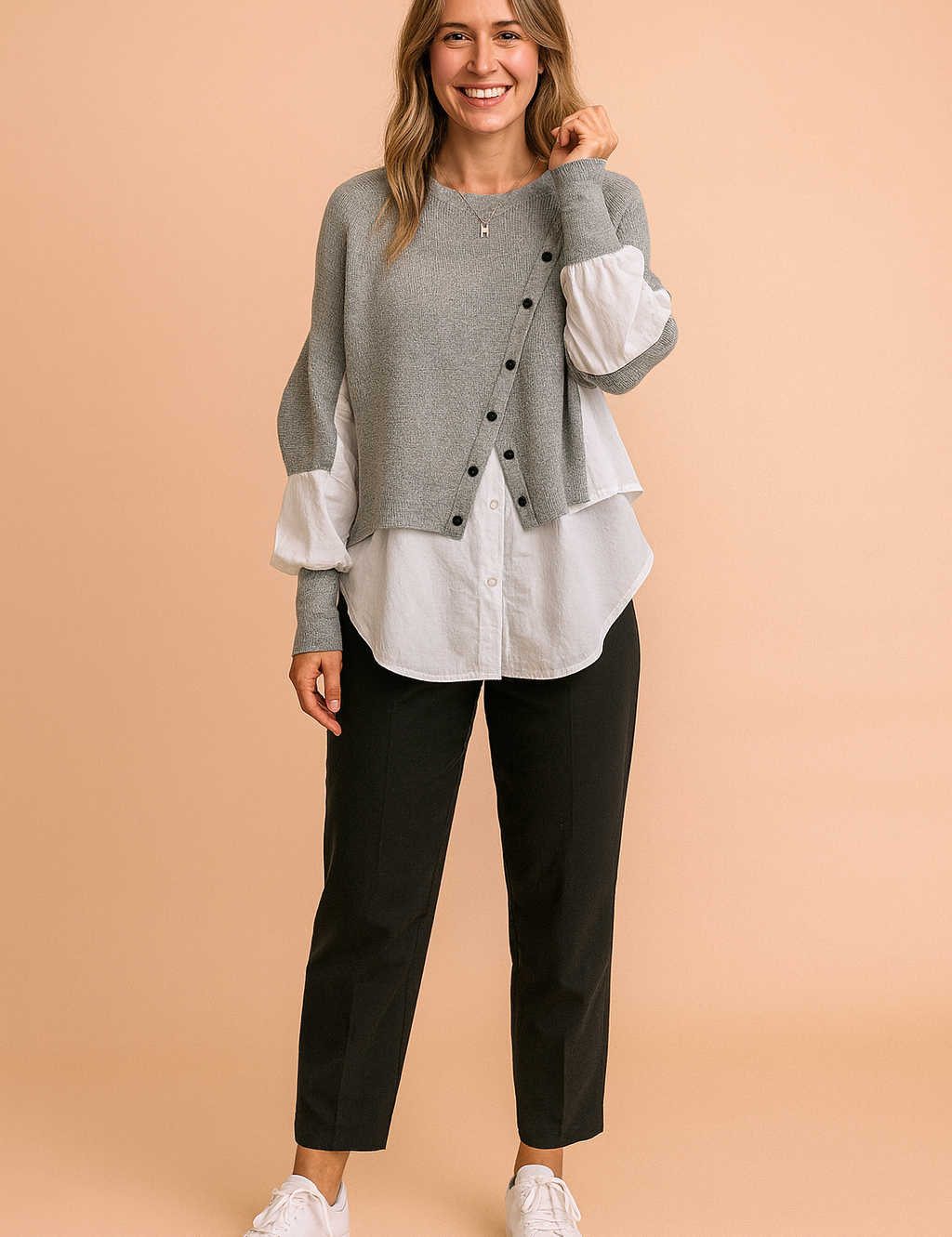 Layered grey button-front knit sweater with asymmetrical design, paired over a crisp white long-sleeve button-down shirt, styled with tailored black trousers and white sneakers.