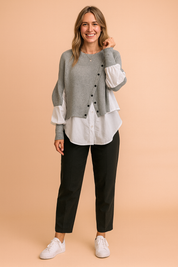 Layered grey button-front knit sweater with asymmetrical design, paired over a crisp white long-sleeve button-down shirt, styled with tailored black trousers and white sneakers.