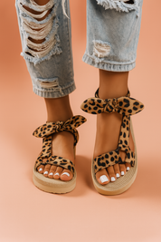 A photograph showcases a pair of women's leopard print platform sandals featuring a T-strap silhouette, soft fabric texture, decorative ankle bows, and beige ridged soles designed for comfort and casual style.