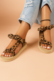  A photograph showcases a pair of women's leopard print platform sandals featuring a T-strap silhouette, soft fabric texture, decorative ankle bows, and beige ridged soles designed for comfort and casual style.