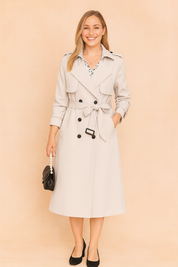 Light beige trench coat with double-breasted button design, belted waist, shoulder epaulets, and mid-calf length, paired with a black handbag and classic black heels.