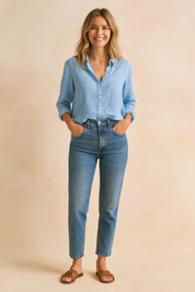 Light blue linen blouse with a relaxed fit, full button-down front, pointed collar, and rolled sleeves, paired with high-rise slim-fit jeans and brown slide sandals for a refined casual look.