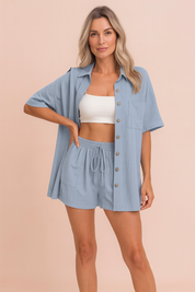 Light blue cotton lounge set with short-sleeved button-down shirt and matching drawstring shorts, styled for summer comfort against a pastel sand pink background.