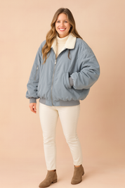 Light gray-blue oversized bomber jacket with white collar lining, styled over a cream turtleneck, white pants, and ankle boots.
