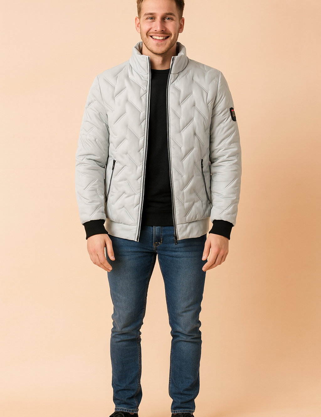 Light grey quilted puffer jacket with diagonal stitching, black ribbed cuffs, high collar, and zippered front pockets, styled with a black crewneck sweater, slim blue jeans, and black suede shoes.