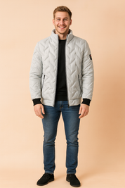 Light grey quilted puffer jacket with diagonal stitching, black ribbed cuffs, high collar, and zippered front pockets, styled with a black crewneck sweater, slim blue jeans, and black suede shoes.