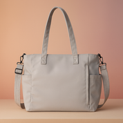 Light grey rectangular tote bag with dual handles, zippered side pocket, and adjustable shoulder strap, displayed on a beige platform against a pastel pink and sand gradient background.
