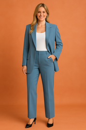 Light steel blue tailored blazer with matching high-waisted straight-leg trousers, styled over a white scoop-neck tank top and paired with black pointed-toe heels, set against an orange sand-tone studio background.