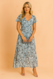 Kobieta w niebieskiej eleganckiej kwiatowej sukience midi Lilia, świetna na codzienne i półformalne letnie stylizacje.