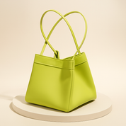 Lime green faux leather handbag with trapezoidal shape and dual handles, featuring smooth texture and clean stitching, displayed on a soft round platform with a gradient neutral background.