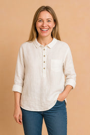 European woman with ash blond hair wearing a crisp white linen shirt and blue jeans, standing against a soft sand pastel background.