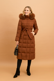 Long quilted brown puffer coat with fur-trimmed hood, belted waist tie, square-stitched pattern, full-length design, and front button closure, paired with black ankle boots and handbag.