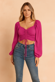 Fashion-forward European woman wearing a vibrant magenta ruched crop top with sheer long sleeves and high-waisted denim jeans, standing confidently against a soft sand and pastel blush background. Ideal for summer fashion editorials and modern casual styling.