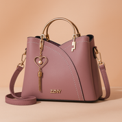Mauve faux leather handbag with gold-tone hardware, heart charm, and stitched detailing, displayed on a beige platform against a pastel blush gradient background.