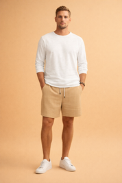 Men’s relaxed summer outfit featuring beige vertically ribbed drawstring shorts with a straight hem, paired with a clean white long-sleeve lightweight shirt and minimal white low-top sneakers, styled in a modern casual silhouette.