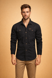 Men’s black long-sleeve button-up shirt with structured collar, front button closure, dual chest flap pockets with buttons, tailored fit, paired with straight-cut beige trousers.
