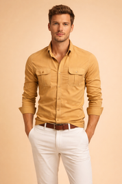 Men’s slim-fit camel sand long-sleeve button-up shirt with structured collar, two symmetrical chest flap pockets, tonal buttons, clean vertical seam lines, and smooth cotton twill fabric, paired with white tailored trousers and a brown leather belt.