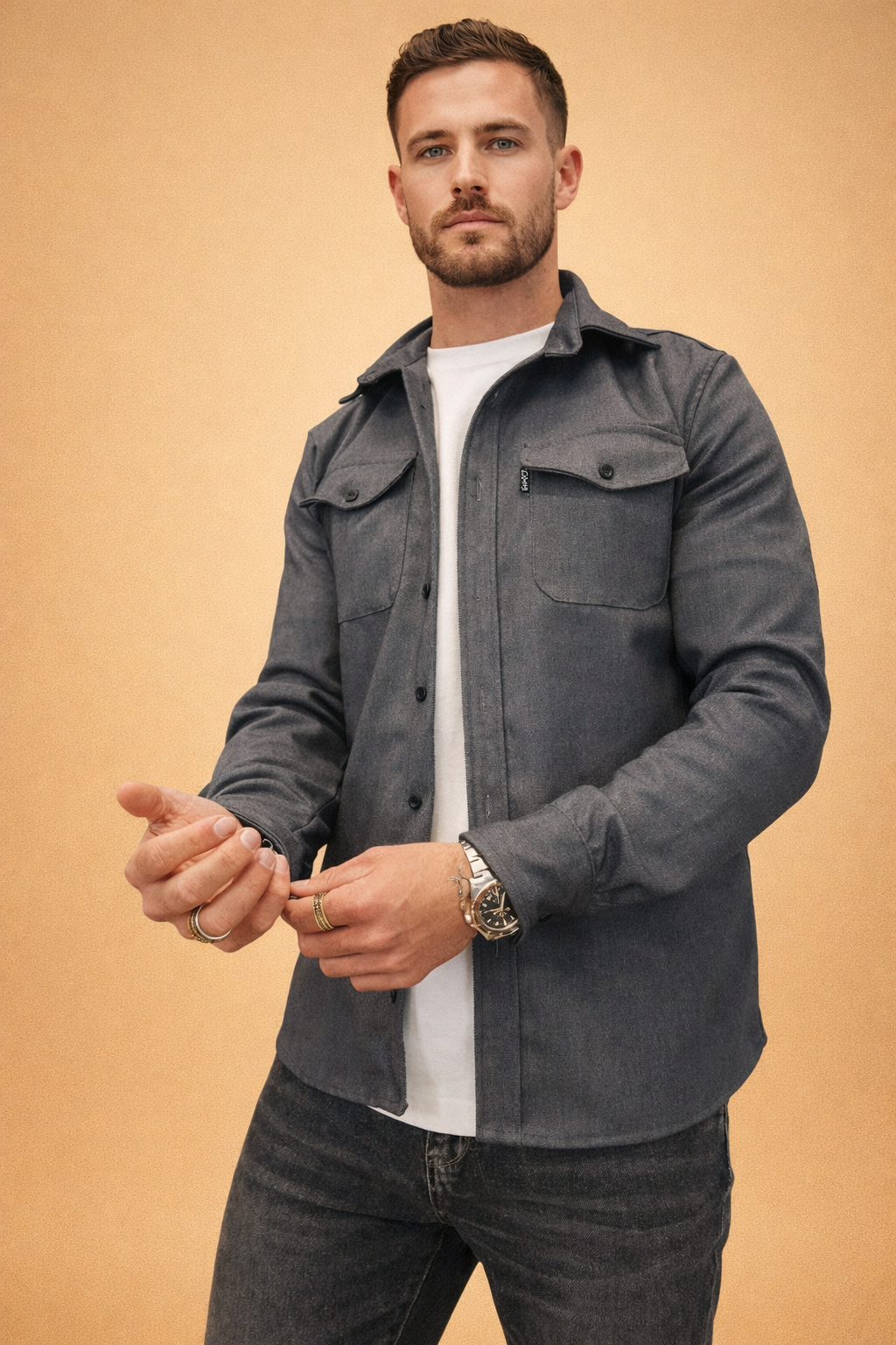 Men’s charcoal grey button-front overshirt with structured collar, dual chest flap pockets, long sleeves with buttoned cuffs, layered over a clean white crew-neck top and styled with slim dark denim trousers