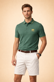 Forest green short-sleeve polo shirt with contrast collar piping and embroidered chest logo, paired with tailored white chino shorts, brown leather belt, and minimalist wristwatch.