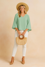 Mint green V-neck blouse with flowing wide sleeves, styled with slim white trousers, straw hat, and a structured woven handbag for a breezy, elegant summer outfit.