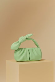 Pastel mint green ruched handbag with bow handle, displayed on a beige pedestal against a soft sand-to-blush gradient background.