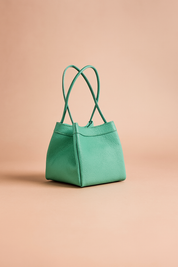 Mint green pebbled leather handbag with structured silhouette and twin looped handles on a pastel gradient background.