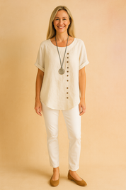 Off-white linen short-sleeve blouse with asymmetrical side button detailing and round neckline, paired with slim white pants and tan flats.