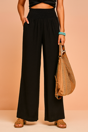 High-waisted wide-leg black smocked pants styled with tan woven sandals and a textured tan leather handbag.