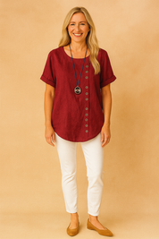Burgundy linen short-sleeve blouse with curved hem and side button design, paired with crisp white fitted pants and tan suede flats.
