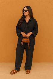 Relaxed-fit black linen button-down shirt with front chest pocket, paired with matching wide-leg black linen trousers, styled with brown leather sandals and a structured tan crossbody bag.