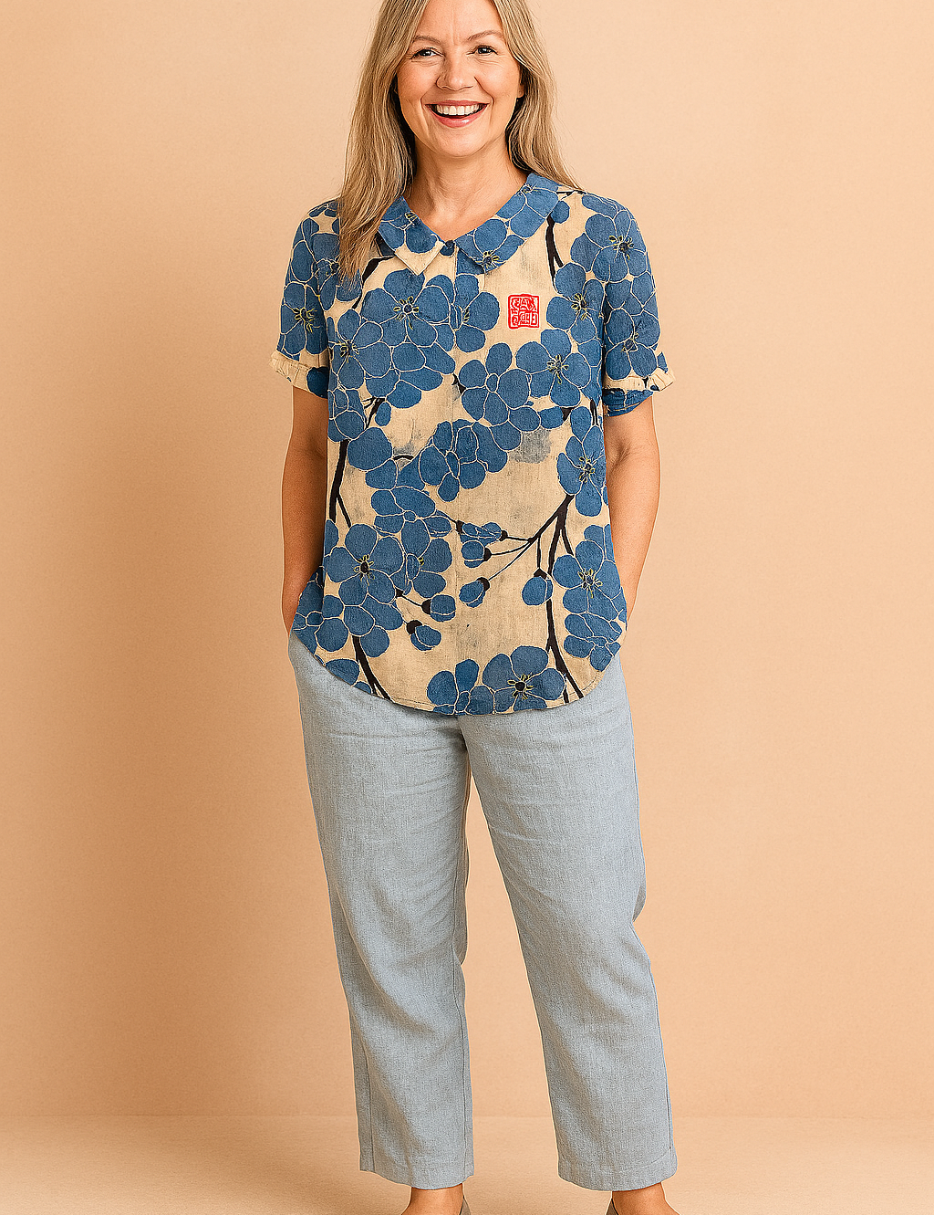 Short-sleeve blouse with a rounded collar featuring oversized indigo-blue floral print and black branch accents on a beige linen-textured background, with a red stamp detail on the chest.