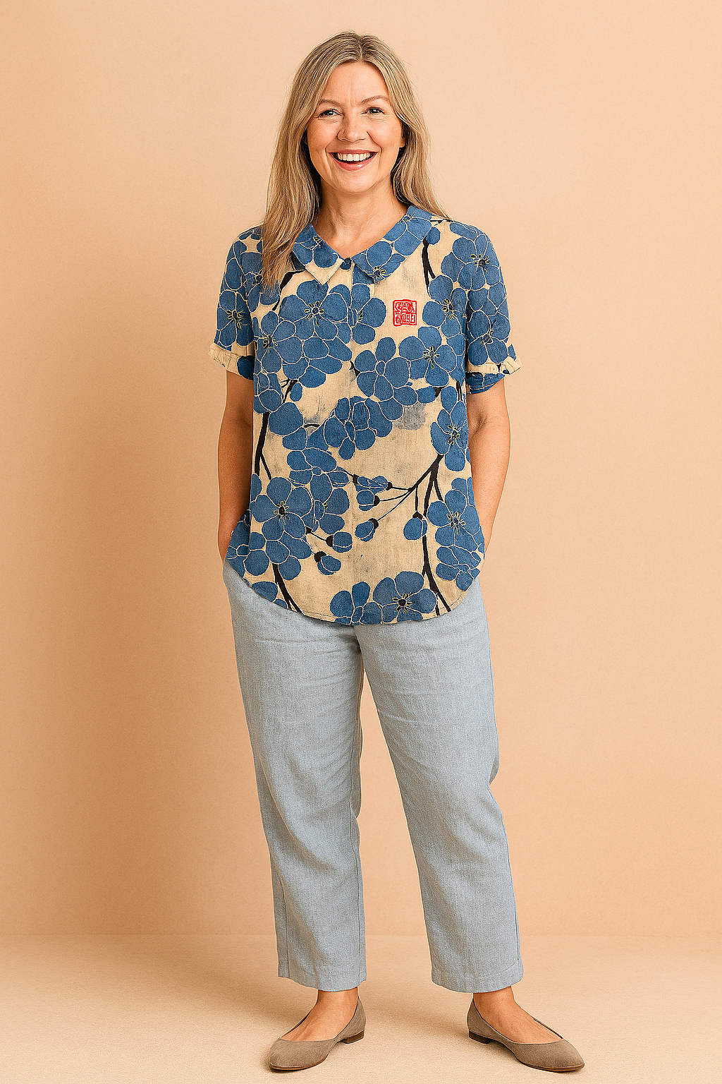 Short-sleeve blouse with a rounded collar featuring oversized indigo-blue floral print and black branch accents on a beige linen-textured background, with a red stamp detail on the chest.