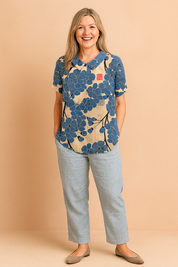 Short-sleeve blouse with a rounded collar featuring oversized indigo-blue floral print and black branch accents on a beige linen-textured background, with a red stamp detail on the chest.