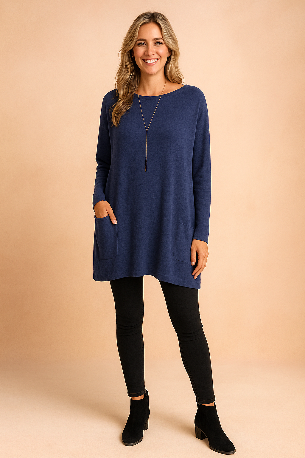Navy blue oversized tunic sweater with long sleeves and dual front pockets, styled with black leggings and suede ankle boots.