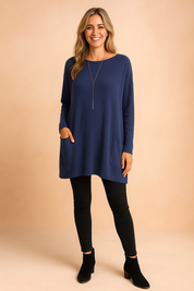 Navy blue oversized tunic sweater with long sleeves and dual front pockets, styled with black leggings and suede ankle boots.