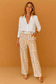 White button-down blouse tucked into high-waisted beige and white geometric print wide-leg pants, accessorized with a white belt, layered jewelry, and beige open-toe sandals.