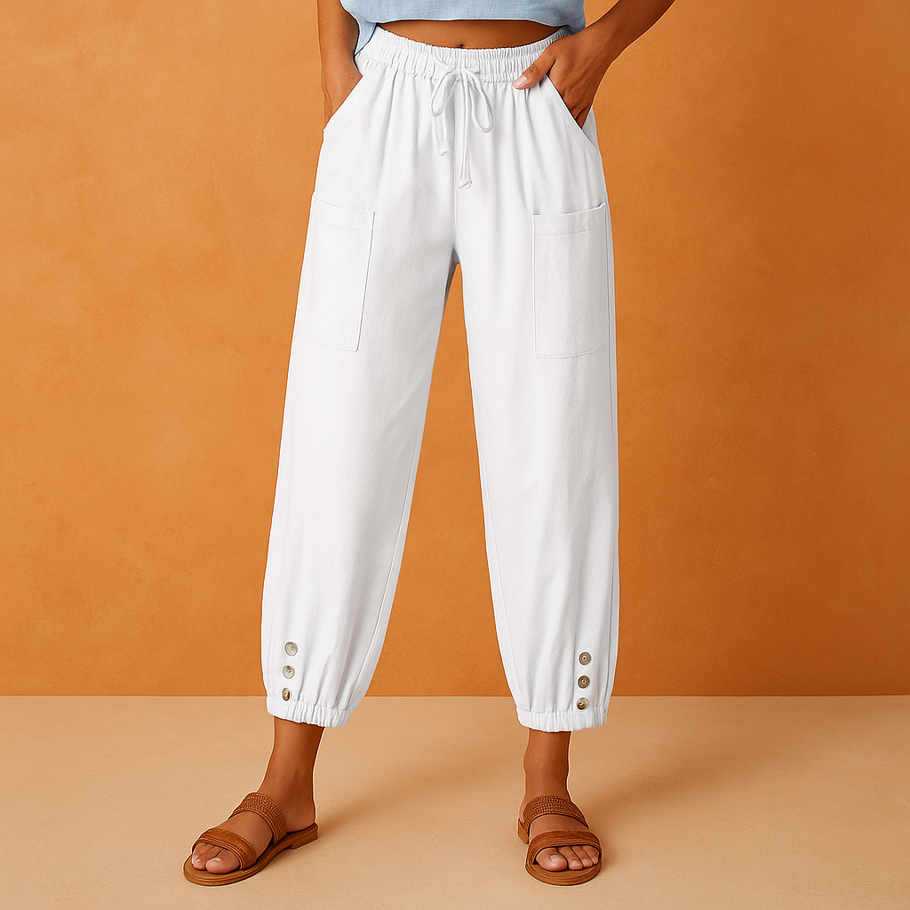 White cropped drawstring pants with large patch pockets and gold button cuffs, styled with tan woven sandals, displayed in a soft orange sand studio setting.
