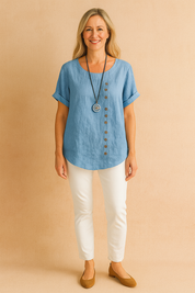 Light blue linen short-sleeve blouse with asymmetrical wooden-style button detailing and curved hem, paired with fitted white pants and tan ballet flats.