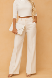 Elegant two-piece outfit featuring a boat-neck three-quarter sleeve crop top paired with high-waisted, wide-leg tailored trousers with sharp front creases, belt loops, and side pockets, completed with open-toe heels and a quilted clutch.