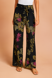 Wide-leg black pants featuring a vivid floral print with olive-green leaves and magenta flowers, styled with a front tie waist and paired with beige sandals.