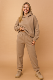 Neutral beige loungewear set featuring a relaxed-fit hoodie with dropped shoulders and matching high-waisted jogger pants made from soft fleece fabric, styled with beige platform sneakers.


