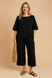Black linen two-piece set featuring a relaxed-fit elbow-sleeve top and matching cropped pants with a folded hem, styled with black sandals and a woven straw handbag.